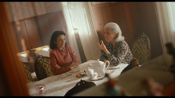 Dos mujeres sentadas en una mesa tomando té. Una de cabello oscuro sonríe a otra de cabello blanco que come un pequeño dulce