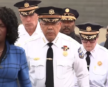 El comisionado Kevin Bethel y otros oficiales de policía caminan con uniforme de gala blanco y gorras ceremoniales
