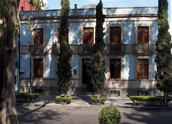 Edificio azul claro de dos pisos con ventanas de madera y balcones de hierro. Cuatro cipreses altos y arbustos frente a la fachada, con sombras en la calle