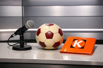 Un micrófono negro, una pelota de fútbol crema y burdeos, y una carpeta naranja con la letra K blanca en un logo cuadrado, sobre una mesa en un estudio.