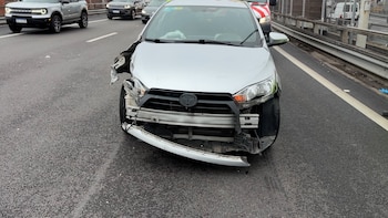 Automóvil plateado con el parachoques y la parte frontal dañados, en una carretera asfaltada. Otros vehículos y una barandilla metálica son visibles