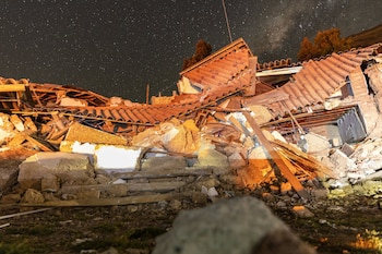 Restos de una casa derrumbada con techo de tejas, vigas de madera y escombros de mampostería bajo un cielo nocturno con estrellas y Vía Láctea