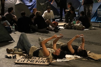 Dos jóvenes acostados en el suelo usan sus teléfonos junto a un cartel de huelga general y varias tiendas de campaña