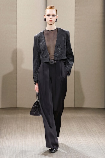 Una mujer con cabello rojizo liso, blusa transparente, chaqueta negra estampada, pantalón negro ancho, cinturón, cartera negra y zapatos negros camina sobre un piso claro