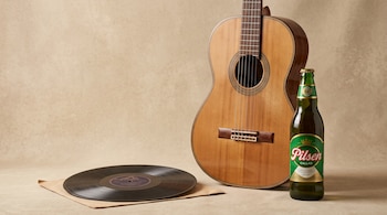 Una guitarra de madera, un disco de vinilo negro y una botella de cerveza Pilsen Callao sobre un fondo beige.