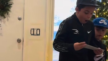 Un niño con gorra negra y chaqueta oscura, con la boca abierta de asombro, sostiene un papel mientras otro niño con gorra azul mira detrás de él
