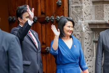 Keiko Fujimori sonríe y saluda, vestida de azul, junto a un hombre de traje negro y corbata roja que también saluda, frente a una puerta de madera