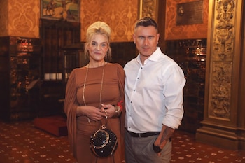 Una mujer rubia y un hombre de pelo oscuro posan en un interior con paredes doradas y ornamentadas. La mujer, vestida de marrón, sostiene un bolso redondo negro brillante