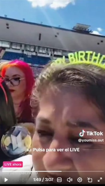 Una mujer lleva una diadema con la leyenda Happy Birthday en un estadio, mientras otra persona al lado sostiene un bastón de luz de BTS