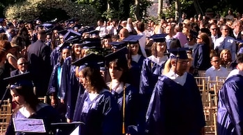 Estudiantes vistiendo togas y birretes azules caminan frente a invitados durante una ceremonia de graduación al aire libre
