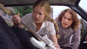 Primer plano de Julia Stiles y Heath Ledger dentro de un coche. Stiles sujeta una guitarra eléctrica blanca, mientras Ledger, con pelo rizado, la mira sonriendo