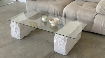 Mesa de centro con tapa de cristal transparente y dos bases de piedra blanca rugosa, con una vela y un cuenco de guijarros. Un sofá beige se ve al fondo.