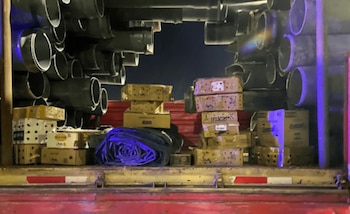 Interior de un camión que muestra grandes tubos grises y pilas de cajas de cartón perforadas. Una lona azul enrollada se ve en el centro. El entorno es oscuro