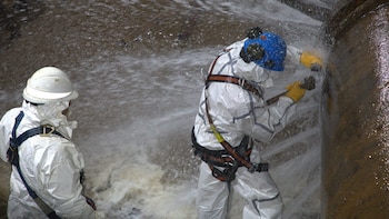 Dos personas con trajes blancos, cascos y arneses manipulan una tubería metálica de la que sale agua a presión