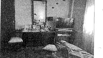 Imagen en blanco y negro de un dormitorio con cama, tocador con fotos, televisor y una ventana con cortinas a la derecha