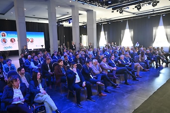 Personas sentadas en filas de sillas dentro de un salón iluminado con luces azules frente a un escenario con pantalla