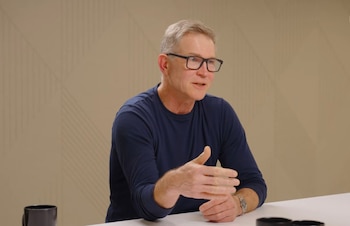 Un hombre caucásico de mediana edad con cabello gris, gafas negras y camiseta azul marino, sentado en una mesa blanca, gesticula con la mano derecha