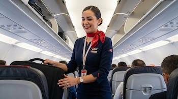 Una tripulante de cabina de Avianca sonríe mientras guarda un equipaje de mano negro en el compartimento superior de un avión, con pasajeros sentados detrás.