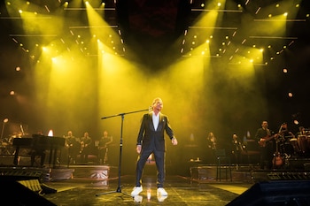El cantante Ricardo Montaner en el escenario durante un concierto, vistiendo un traje oscuro y zapatillas blancas, bajo intensas luces amarillas y humo