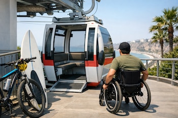 Persona en silla de ruedas en estación de teleférico con rampa de acceso, junto a cabina abierta, bicicletas y tabla de surf. Playa y ciudad costera al fondo.