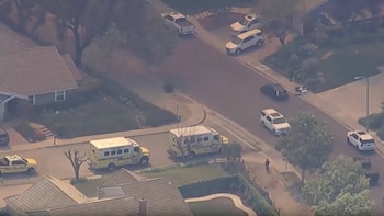 Vista aérea de una calle residencial con dos vehículos de emergencia amarillos, varios coches aparcados y uno en movimiento, casas y un peatón en la acera
