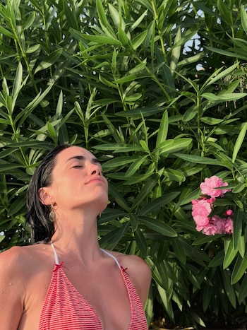 Mujer de cabello oscuro y mojado, con un bikini de rayas rojas y blancas, cierra los ojos bajo el sol frente a un follaje verde con flores rosadas
