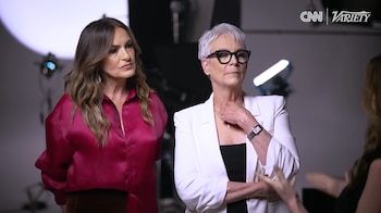 Dos mujeres en un estudio. Una con blusa fucsia y pelo castaño largo; la otra con blazer blanco, pelo gris corto y gafas. Fondo con luces de estudio