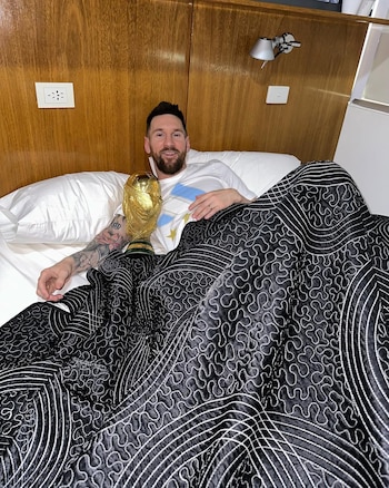 Lionel Messi, con barba y camiseta blanca, sonríe acostado en una cama con sábanas blancas, cubierto con un edredón negro, junto a la Copa del Mundo dorada