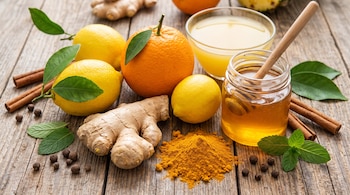 Naranjas, limones, raíz de jengibre, cúrcuma en polvo, canela y un frasco de miel sobre una mesa de madera.