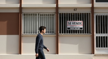 Un joven camina frente a la fachada de un edificio color crema y marrón con ventanas enrejadas. Se observa un cartel de "Se Vende" en una ventana.