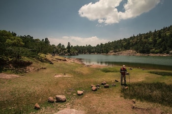 Un hombre con gorra, camisa a cuadros y bastón de senderismo está de pie en la orilla de una laguna. El paisaje incluye árboles, rocas y cielo con nubes