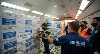 Interior de avión de carga con palés de cajas que dicen "SARAMPIÓN VACUNA" y "GOBIERNO DE GUATEMALA", un cartel que indica "2,010,000,000 DOSIS", y hombres con uniformes.