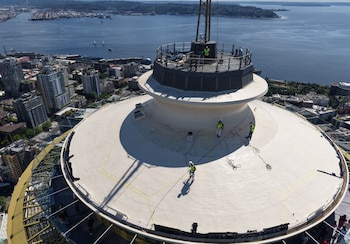 Vista aérea de trabajadores con equipo de seguridad realizando construcción en la cubierta superior de la Aguja Espacial, con la ciudad de Seattle y el agua de fondo