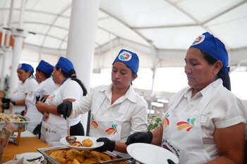 Una mujer sonriente con gorro y delantal blanco con un logo de comida, sostiene una bandeja negra con tres platos de comida. Al fondo, personas y pancartas