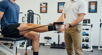 Primer plano de un fisioterapeuta ayudando a un deportista sentado con un ejercicio de banda de resistencia para su pie y tobillo lesionados.