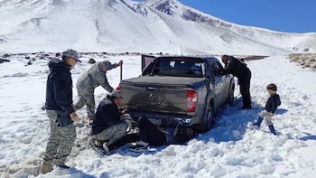 Un grupo de hombres y un niño permanecen junto a una camioneta gris varada en un camino nevado frente a montañas