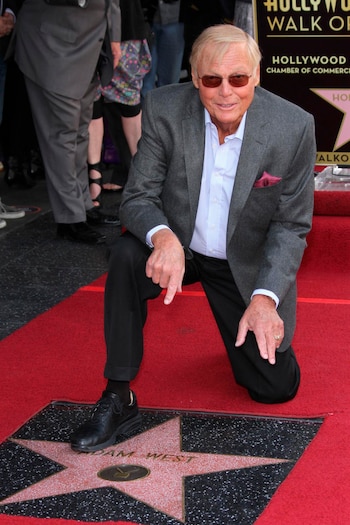 Adam West, un hombre mayor con gafas de sol oscuras, blazer gris y camisa a rayas azul y blanca, se arrodilla en una alfombra roja señalando su estrella