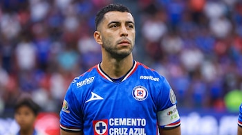 Un jugador de fútbol con camiseta azul de Cruz Azul mira hacia adelante en un estadio con el público al fondo.