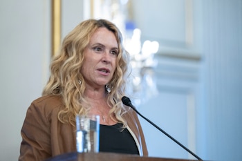 Mujer de mediana edad con cabello rubio rizado y chaqueta marrón, hablando en un micrófono situado en un podio, con un vaso de agua