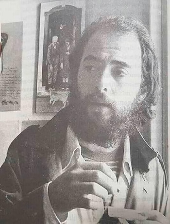 Fotografía en blanco y negro de El Indio Solari con barba y cabello largo, mirando fijamente a la cámara, mientras sostiene un objeto pequeño