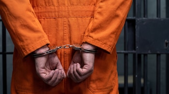 Primer plano de las manos de una persona esposadas por la espalda, vestida con un uniforme de color naranja, frente a barras de una celda.