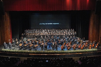 Un escenario con cortinas rojas y un gran coro, una orquesta con instrumentos de cuerda y viento, y cuatro solistas al frente. Pantalla con texto y público en butacas