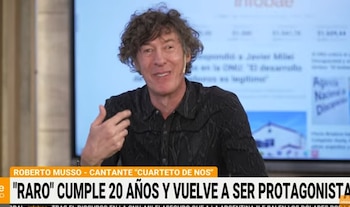 Un hombre de cabello rizado y camisa negra gesticula con la mano frente a una pantalla de televisión en un estudio