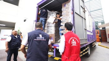 Varias personas con chalecos de ONPE y JNE cargan cajas de material electoral en un camión morado y blanco. Un hombre mira hacia arriba en primer plano