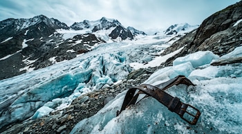 Una correa de cuero con hebilla metálica sobre hielo glaciar en primer plano, con montañas rocosas y nevadas en el fondo.