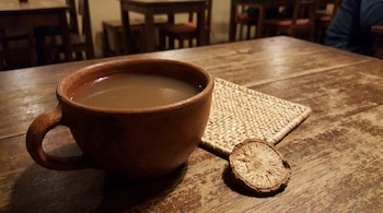 Una taza de cerámica marrón con un líquido color café claro sobre una mesa de madera, junto a una esterilla tejida y una rodaja de raíz seca.