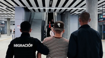 Hombres de espaldas en un interior moderno. Uno usa chaleco "MIGRACIÓN", otro camiseta a rayas y gorra. Al fondo se ven escaleras y otros peatones.