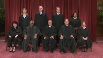 Nueve jueces con togas negras posan para una foto grupal. Cinco hombres y cuatro mujeres sentados y de pie, con un telón rojo de fondo