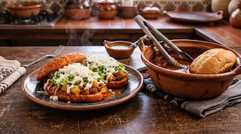 Plato con dos pambazos rellenos junto a una cazuela de barro con salsa, pinzas metálicas y un bolillo sobre un paño de tela.