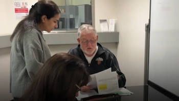 Un hombre con gafas sentado y una mujer de pie revisan documentos en una mesa de oficina, con una tercera persona sentada y parcialmente visible